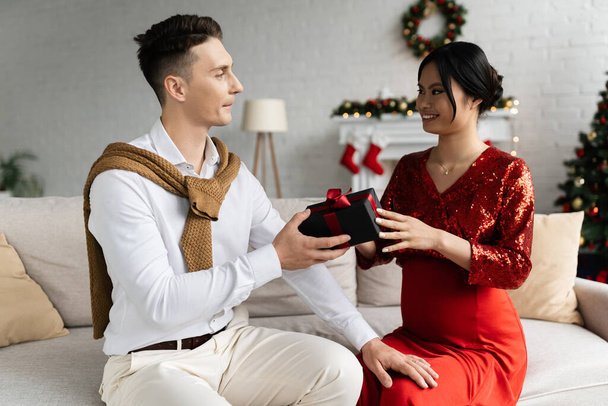 young man presenting Christmas gift to pregnant and elegant asian wife while sitting on couch at home - Foto, immagini