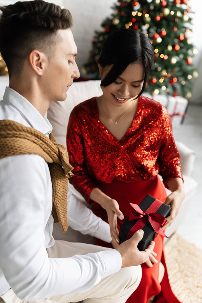 Genç koca, evdeki kanepede hamile Asyalı karısına Noel hediyesi veriyor. - Fotoğraf, Görsel
