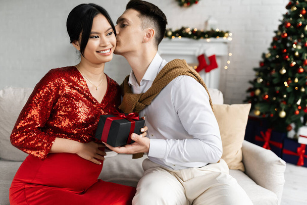 έγκυος Ασιάτισσα με κομψά ρούχα χαμογελώντας δίπλα στον σύζυγο με δώρο Χριστουγέννων να τη φιλάει στον καναπέ στο σπίτι - Φωτογραφία, εικόνα