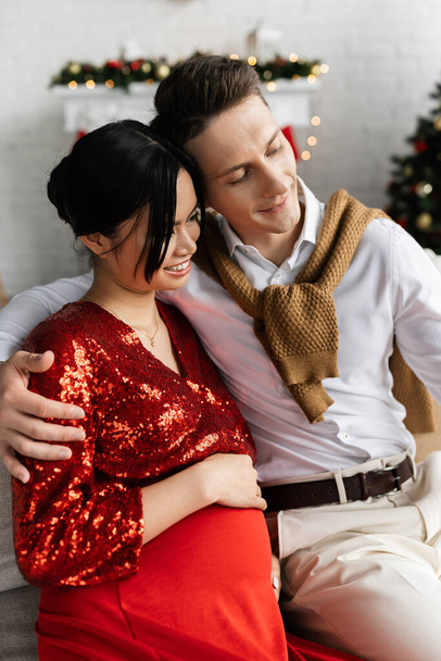 pleased man embracing pregnant asian wife in red and shiny clothes while celebrating Christmas at home - Photo, Image