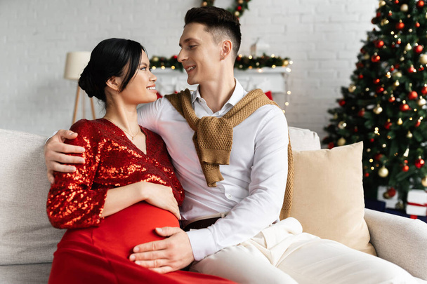 embarazada asiático mujer y marido en festivo ropa mirando uno al otro en sofá cerca borrosa árbol de Navidad - Foto, Imagen