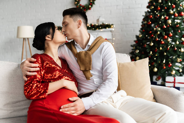 man kissing pregnant asian woman and embracing her tummy on couch near blurred Christmas tree - 写真・画像