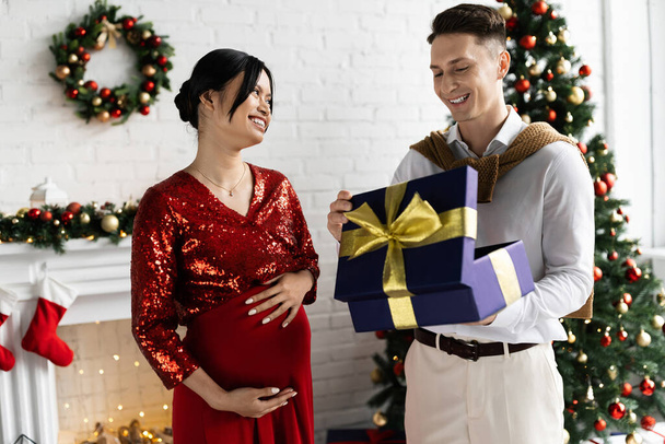 pregnant asian woman smiling near happy husband opening gift box in living room with Christmas decor - Φωτογραφία, εικόνα