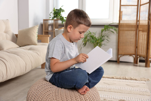 Boy with poor posture reading book on beige pouf in living room. Symptom of scoliosis - 写真・画像