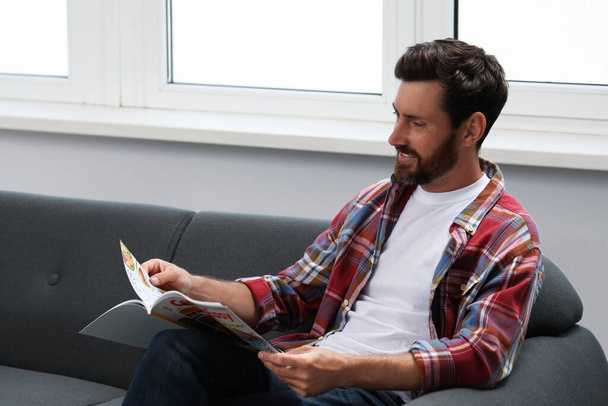 Smiling bearded man reading magazine on sofa indoors - Foto, afbeelding