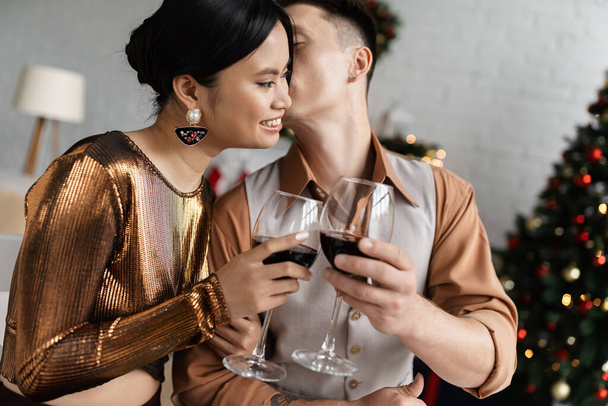 young man kissing happy and elegant asian wife and clinking wine glasses while celebrating Christmas - Фото, зображення