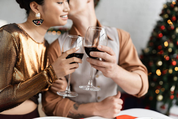 smiling asian woman in shiny blouse clinking wine glasses with blurred husband during Christmas celebration - Photo, image