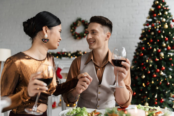 elegante pareja interracial con copas de vino cogidas de la mano y sonriéndose durante la cena romántica de Navidad - Foto, imagen
