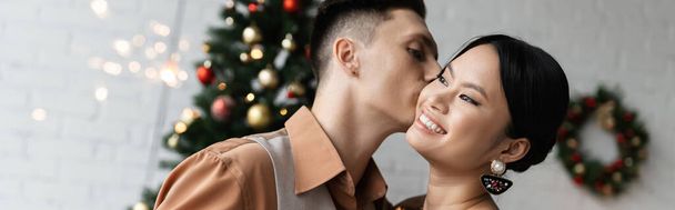young man kissing cheerful asian wife while celebrating Christmas at home, banner - Photo, Image