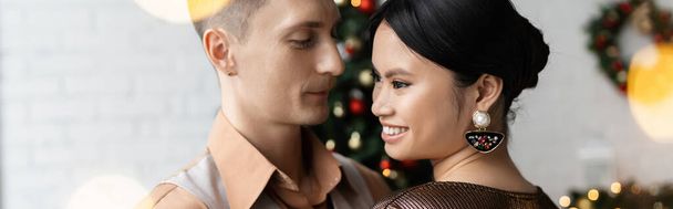 happy man looking at cheerful and elegant asian wife during Christmas celebration, banner - Photo, Image