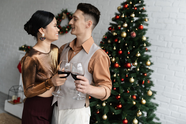 ευτυχισμένο πολυεθνικό ζευγάρι κοιτάζοντας ο ένας τον άλλο κρατώντας ποτήρια κόκκινο κρασί κοντά στο χριστουγεννιάτικο δέντρο  - Φωτογραφία, εικόνα
