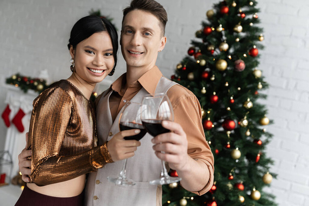heureux couple multiethnique tenant des verres de vin rouge près de l'arbre de Noël  - Photo, image