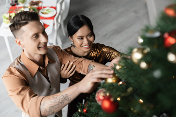 άποψη υψηλής γωνίας χαμογελώντας διαφυλετικό ζευγάρι διακόσμηση χριστουγεννιάτικο δέντρο με λαμπερά μπιχλιμπίδια - Φωτογραφία, εικόνα