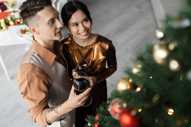 visão de alto ângulo de casal interracial feliz em roupas festivas segurando copos de vinho perto da árvore de Natal  - Foto, Imagem