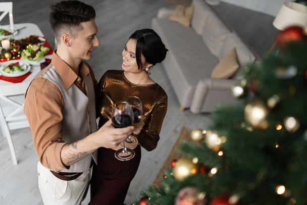 Blick aus der Vogelperspektive auf ein glückliches interrassisches Paar in stylischem Outfit, das in der Nähe des Weihnachtsbaums Weingläser klappert  - Foto, Bild