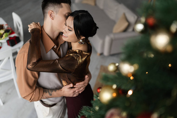 alto ângulo vista de feliz asiático mulher abraçando tatuado marido perto decorado Natal árvore  - Foto, Imagem
