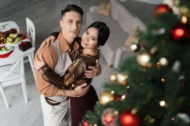 high angle view of cheerful asian woman hugging with husband near Christmas tree  - Zdjęcie, obraz