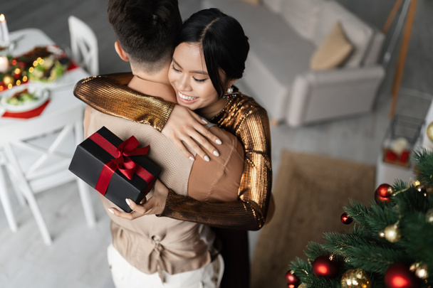 high angle view of cheerful asian woman hugging husband while holding Christmas present  - Fotoğraf, Görsel