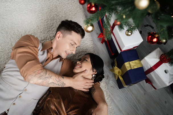 top view of cheerful interracial couple lying under Christmas tree with presents  - Photo, Image