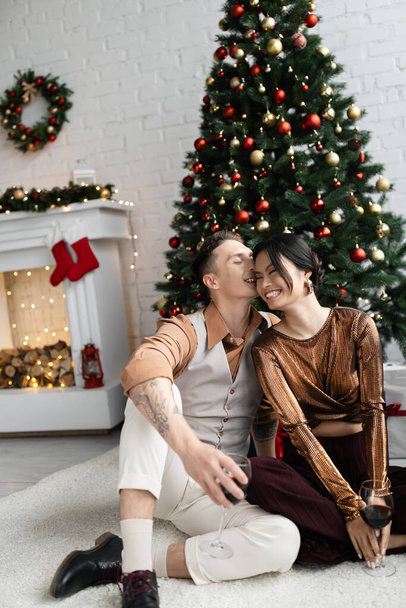 alegre homem beijar bochecha de feliz asiático esposa enquanto sentado sob Natal árvore  - Foto, Imagem