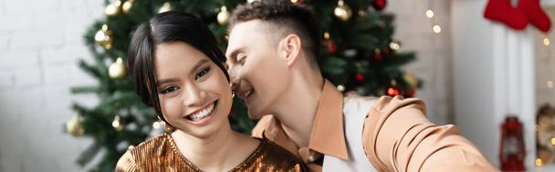 homme gai chuchotant à l'oreille de heureuse femme asiatique près de l'arbre de Noël, bannière - Photo, image