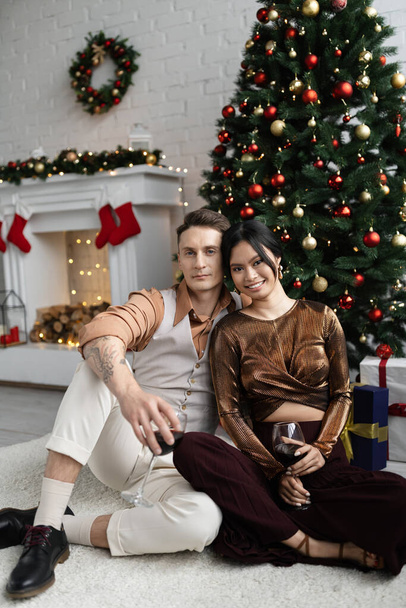 cheerful man and happy asian woman holding glasses of wine while sitting under Christmas tree  - Foto, imagen