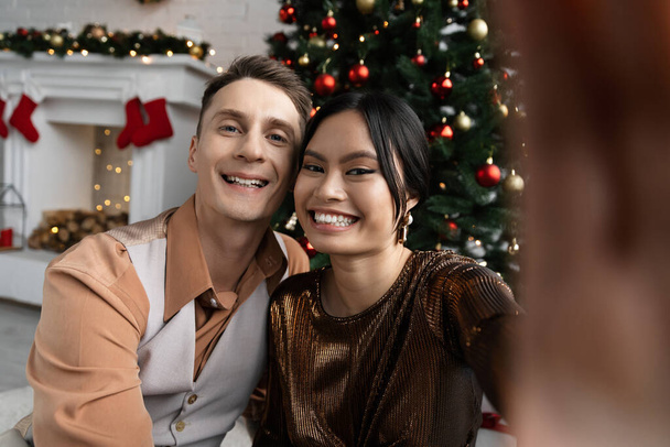 happy interracial couple smiling and looking at camera while sitting under Christmas tree  - 写真・画像