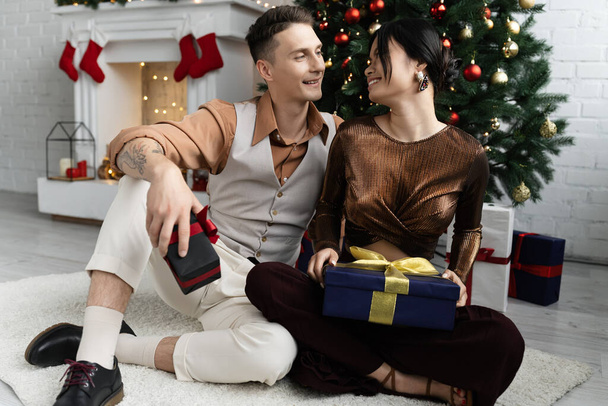 heureux couple interracial souriant et tenant cadeaux de Noël tout en étant assis sous le pin décoré  - Photo, image