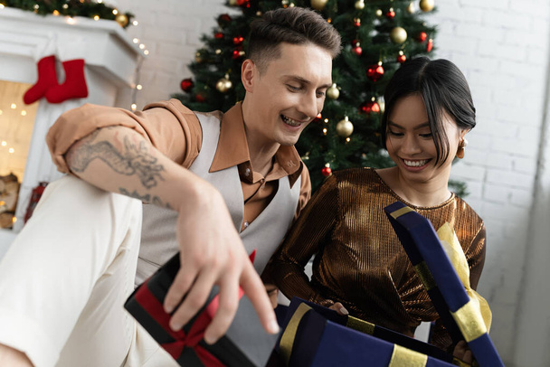happy interracial couple opening Christmas present while sitting under decorated fir  - 写真・画像