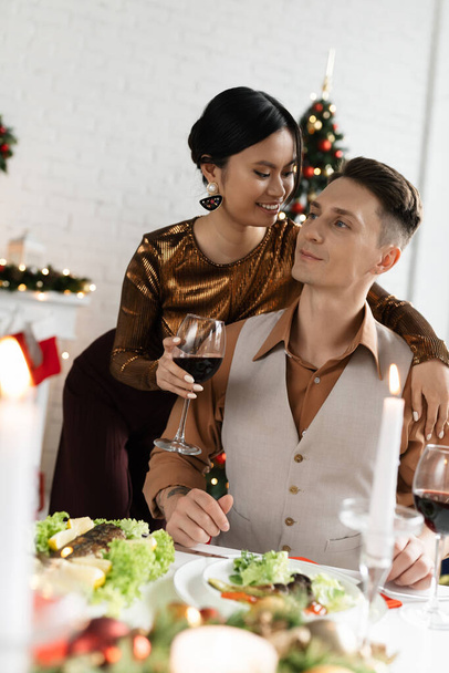 χαρούμενη Ασιάτισσα που κρατάει ένα ποτήρι κρασί και αγκαλιάζει τον άντρα της κοντά στο εορταστικό δείπνο κατά τη διάρκεια των Χριστουγέννων  - Φωτογραφία, εικόνα
