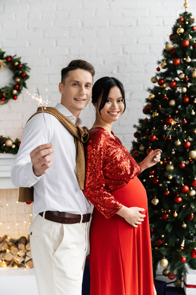 Χαρούμενος άντρας που κρατάει σπίθες με την έγκυο Ασιάτισσα γυναίκα του κοντά στο χριστουγεννιάτικο δέντρο  - Φωτογραφία, εικόνα