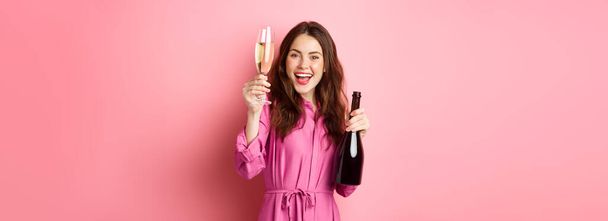 Концепция праздников и праздников. Красивая женщина произносит тост, поднимает бокал шампанского и держит бутылку, веселится на вечеринке, стоя на розовом фоне. - Фото, изображение