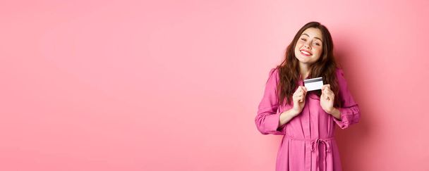 Young brunette woman hugging her credit card with delight, smiling pleased, standing against pink background. Copy space - Фото, изображение