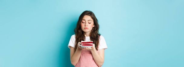 Feestdagen en feestdagen. Triest schattig meisje vieren haar verjaardag alleen, met taart op bord met een kaars en mokken boos, staan eenzaam tegen een blauwe achtergrond. - Foto, afbeelding