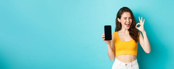 Atraktivní moderní žena v letních šatech, jiskřivě mrká, ukazuje v pořádku nápis s prázdným displejem smartphonu, ukazuje vaše logo na displeji, modré pozadí. - Fotografie, Obrázek