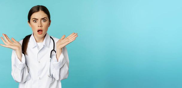 Portrait of shocked female doctor, worried healtcare worker in white robe, looking concerned at patient, standing over blue background. - Фото, изображение