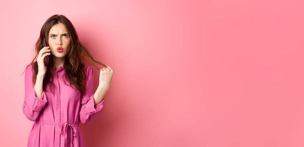 Νεαρή γυναίκα απαντά στο τηλεφώνημα, αναζητούν σύγχυση και αναποφάσιστη, καθιστώντας παραγγελία takeaway στο smartphone, στέκεται σε ροζ φόντο. - Φωτογραφία, εικόνα