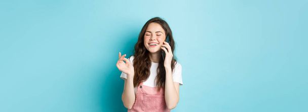 Porträt eines netten und stilvollen Mädchens, das lacht, während es telefoniert, das Smartphone ans Ohr hält und glücklich lächelt, lässig Gespräche führt, vor blauem Hintergrund steht. - Foto, Bild