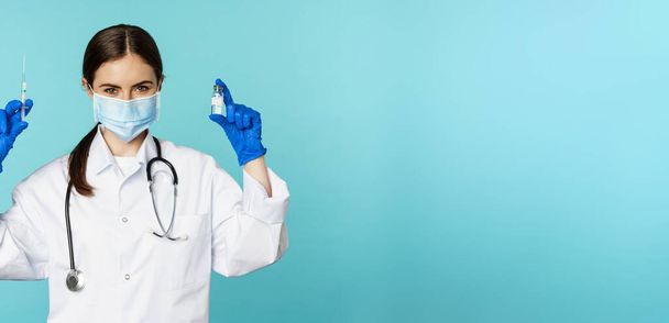 笑顔の医療スタッフ、顔のマスクとゴム手袋の医者、 covid-19パンデミックから注射器とワクチンを示し、青い背景の上に立って. - 写真・画像