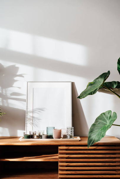 Modern minimalist Scandinavian style interior with white poster mockup, candles and tropical green home plants on a wooden console under sunlight and shadows on a gray wall. Selective focus. - Foto, afbeelding