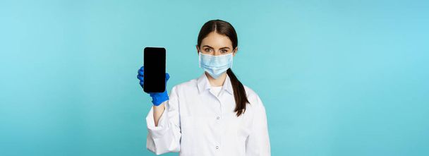 Young woman doctor in medical face mask and hospital uniform, showing mobile phone app, screen interface, online clinic concept, standing over torquoise background. - 写真・画像