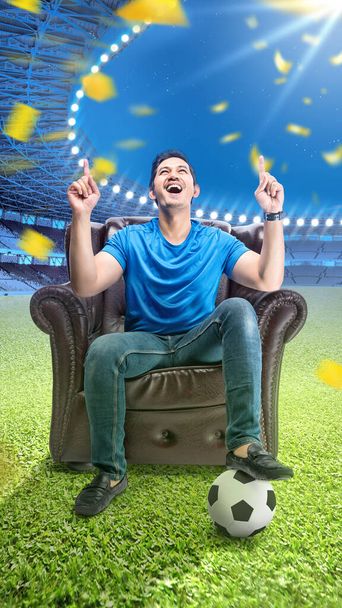 asiatico uomo con il palla seduta su il divano con un eccitato espressione in il calcio stadio sfondo - Foto, immagini