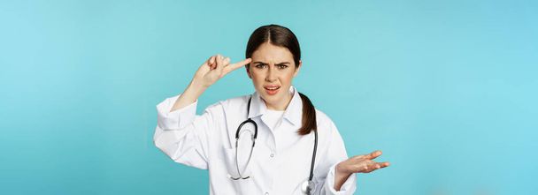 Médico mujer molesta señalando con el dedo a la cabeza, regañando a alguien estúpido, loco o extraño, de pie sobre fondo de color turquesa. - Foto, imagen
