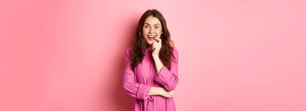 Beautiful excited girl smiling, biting finger and looking with tempted face at camera, having interesting idea, looking at something intriguing, standing against pink background. Copy space - Fotoğraf, Görsel