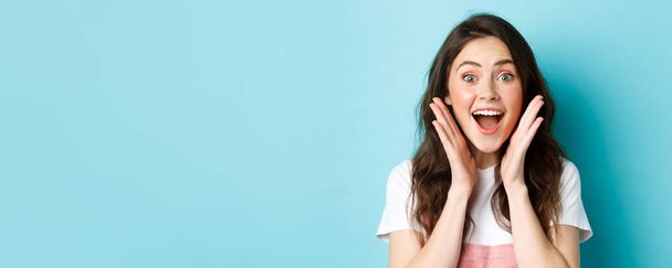 Close up of excited young woman holding hands near face, gasping and looking amazed at camera, stare surprised at super cool news, standing over blue background. - Photo, Image