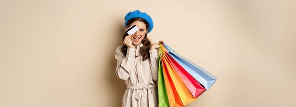 Portrat of trendy feminine girl posing with shopping bags from store and credit card, paying contactless, buying with discount on sale, beige background. - Zdjęcie, obraz