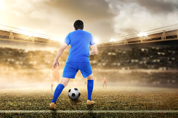 Rear view of Asian football player man in a blue jersey dribbling the ball on the football field - Φωτογραφία, εικόνα
