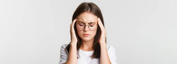 Close-up van jonge vrouw in glazen die het hoofd aanraken en grimmig worden van pijn, hoofdpijn of migraine, misselijkheid. - Foto, afbeelding