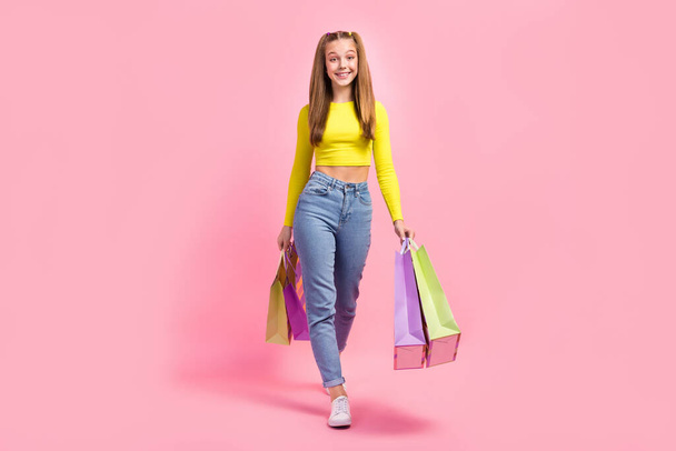 Full length photo of pretty sweet school girl dressed yellow crop top walking holding bargains isolated pink color background. - Foto, Imagen