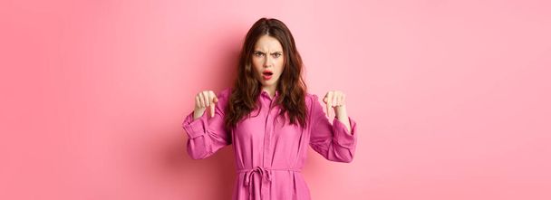 Mujer joven enojada y disgustada frunciendo el ceño, jadeando ofendido, señalando con los dedos hacia abajo, de pie sobre el fondo rosa. - Foto, Imagen